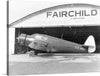 This black and white photograph captures the timeless beauty of the Fairchild JK-1, a small, propeller-driven aircraft that was developed by Fairchild Aviation in the early 1900s. The aircraft is shown parked outside a Fairchild Airplanes hangar, with its sleek lines and elegant design on full display.