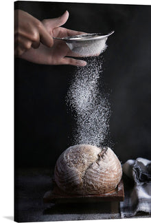  Capture the timeless art of bread making in a single mesmerizing moment with this exquisite print. Against a dark, dramatic backdrop, a hand delicately dusts a freshly baked loaf of bread with flour. 