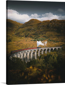  Immerse yourself in the enchanting landscape captured in this print, where nature and engineering unite harmoniously. A majestic stone viaduct, reminiscent of iconic scenes from beloved tales, stretches across the lush greenery of rolling hills. A train, a symbol of journey and discovery, traverses this architectural marvel amidst a backdrop of towering mountains veiled by the soft embrace of clouds. 