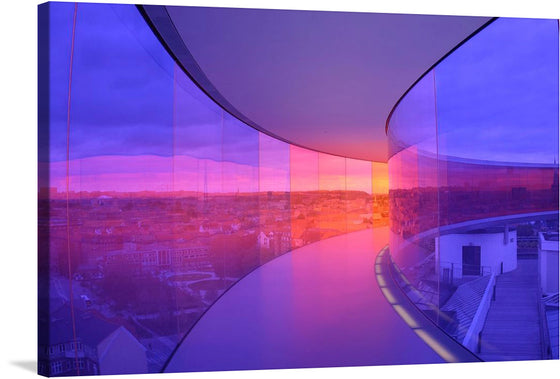 This photograph captures the mesmerizing beauty of a city sunset from a unique perspective. The scene unfolds through the curved glass windows of a modern building, framing the vibrant hues of the sky and the cityscape below. The sunset, with its radiant orange and pink colors, takes center stage, while the city buildings add depth to the scene.