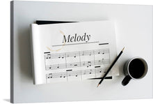  Immerse yourself in the serene elegance of our latest art print, “Melody.” This exquisite piece captures a moment of peaceful reflection, where a sheet of music rests atop a blank canvas journal, accompanied by a pencil and a cup of coffee. The gentle stain on the musical score adds an element of lived experience, evoking feelings of nostalgia and the timeless beauty of music. 