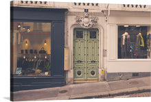  “Paris” is a captivating artwork that encapsulates the city’s timeless elegance and modern charm. The print features a street view in Paris featuring two boutique storefronts flanking an ornate green door. The left boutique has a dark facade with large glass windows showcasing men’s clothing, bags, and hats illuminated by hanging lights. 