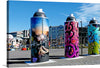 “Paint Cans Three” by Bernard Spragg. NZ is a vibrant and eclectic artwork that encapsulates the dynamic fusion of street art and classic artistry. The image features three oversized paint cans as public art installations, each adorned with different styles of graffiti. The first can on the left displays a dark theme with hands reaching out towards light and sparkles; it has signatures including “WILSON”. 