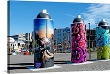  “Paint Cans Three” by Bernard Spragg. NZ is a vibrant and eclectic artwork that encapsulates the dynamic fusion of street art and classic artistry. The image features three oversized paint cans as public art installations, each adorned with different styles of graffiti. The first can on the left displays a dark theme with hands reaching out towards light and sparkles; it has signatures including “WILSON”. 