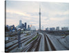 This photograph captures the iconic CN Tower in Toronto, Canada, from a unique perspective. The image foregrounds train tracks, leading the viewer’s eye towards the tower standing majestically in the background. The light blue sky, scattered with clouds, adds depth to the scene.