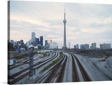  This photograph captures the iconic CN Tower in Toronto, Canada, from a unique perspective. The image foregrounds train tracks, leading the viewer’s eye towards the tower standing majestically in the background. The light blue sky, scattered with clouds, adds depth to the scene.