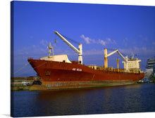  The “Ship AS Vega” print captures the majestic presence of a large, red and white cargo ship docked gracefully against the backdrop of a serene blue sky. The ship, marked with the name “AS VEGA,” is equipped with towering cranes, ready to manage hefty loads. The reflection of the setting sun casts a golden hue on parts of the ship and water, adding a touch of warmth to this industrial scene. 