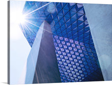  This print is a stunning architectural photograph of a modern building with a unique geometric design. The blue sky and sunburst add a touch of natural beauty to the image, making it a perfect addition to any home or office. The photo-realistic image showcases a modern building with triangular panels in different shades of blue, framed by two concrete pillars on the left and right side. The sky is a bright blue with a sunburst in the top left corner.