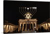 The image features the iconic Brandenburg Gate with a large menorah. The image is a realistic photograph of the Brandenburg Gate in Berlin at night. The gate is illuminated with warm yellow lights. In front of the gate is a large menorah with lit candles.