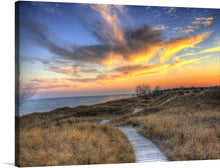 “Colorful Dusk Above The Dunes” is a beautiful print that captures the essence of a peaceful evening at the beach. The sky is a vibrant orange and yellow with wispy clouds. The landscape consists of sand dunes with tall grasses and a wooden walkway.