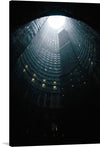 “Johannesburg, South Africa” is a mesmerizing artwork that captures the architectural majesty and mystery of the city’s iconic Ponte City Apartments. The artwork features an upward view inside a cylindrical building structure, taken at night or in low light conditions. The top of the cylinder is open, allowing natural light to illuminate part of the interior. 