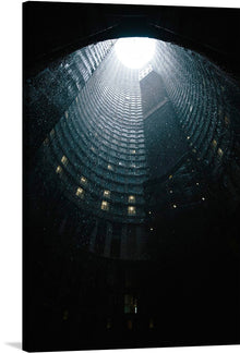  “Johannesburg, South Africa” is a mesmerizing artwork that captures the architectural majesty and mystery of the city’s iconic Ponte City Apartments. The artwork features an upward view inside a cylindrical building structure, taken at night or in low light conditions. The top of the cylinder is open, allowing natural light to illuminate part of the interior. 