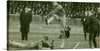 This captivating print captures a riveting moment from an early 20th-century football match. The black and white image is alive with action, featuring players in dynamic movement, their intensity indicative of the high stakes of the game. Spectators, dressed in period attire, add to the historical ambiance, their rapt attention underscoring the excitement of the event.