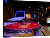 This photograph of a turntable with a red vinyl record and a laptop in a dark room is a stunning and atmospheric image. The red record is the focal point of the photograph, and it is illuminated by a soft light that casts a warm glow over the entire image. The laptop is in the background, and it is barely visible, which helps to create a sense of mystery and intrigue.