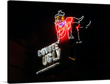  This print captures the essence of a classic neon sign, its vibrant hues and electric glow cutting through the night. The words "Coyote Ugly" are underlined and blaze in white. A lone cowboy girl sits in a glowing red dress and cowboy boots with her leg crossed over the other leg.