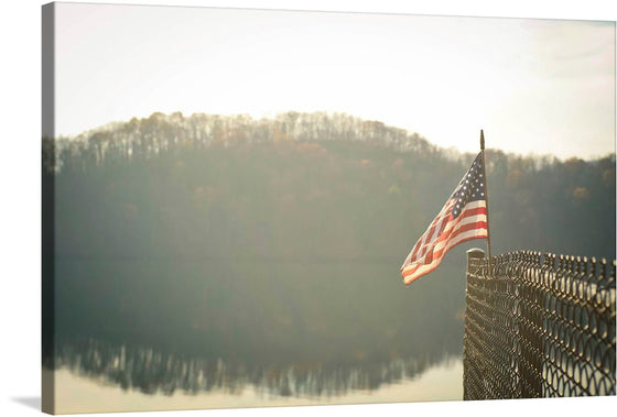 “Raystown Lake, United States” is a captivating print that encapsulates the tranquil ambiance of nature intertwined with national pride. The artwork captures a pristine reflection of the wooded landscape on the calm waters of Raystown Lake, bathed in the soft glow of sunlight. In the foreground, an American flag waves gracefully, symbolizing strength and unity amidst nature’s tranquility. 