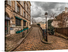  “Paris” is a captivating artwork that transports you to a serene street in the heart of the city. The print features a picturesque scene from a quiet street in Paris, with a cobblestone pathway leading the viewer’s eye through the image. On the left, there’s an old building with signage reading “AU LAPIN AGILE,” indicating it might be a business or café. 