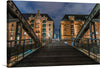 Immerse yourself in the enchanting allure of this exquisite print capturing a serene night at the iconic warehouse district, Speicherstadt in Hamburg. This artwork encapsulates the harmonious blend of historic architecture and modern illumination, offering a visual journey through the wooden bridge that leads to majestic brick buildings adorned with elegant windows.