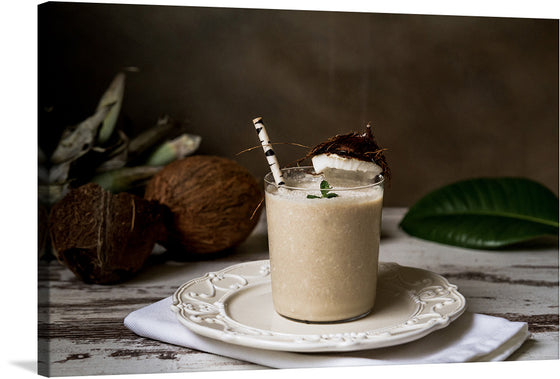 This marvelous photograph of a glass of coconut milk sitting on a plate captures the simple beauty and deliciousness of this tropical beverage. The glass is filled with a milky white liquid, topped with a garnish, and paired with a straw. In the background there is a fresh brown coconut and a green leaf.