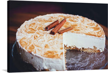  he image is a photo-realistic representation of a cinnamon cheesecake with a slice cut out. The cheesecake is on a glass plate with an intricate design. The top of the cheesecake has a beautiful swirl pattern, and two cinnamon sticks are placed on top of it. 