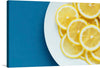 Adorn your space with the vibrant allure of this exquisite art print, where the zesty charm of freshly sliced lemons comes to life. Each slice, rich with the intricate details of pulpy textures and glistening juice, is a testament to nature’s artistry. The lemons rest elegantly on a pristine white plate, their yellow hue gleaming against a backdrop of deep azure - an embodiment of summer’s lively spirit.