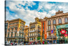 This colorful and captivating print captures the vibrant essence of a bustling Barcelona street. The ornate, colorful buildings stand tall against a clear blue sky, their intricate details inviting you to explore every nook and cranny. As sunlight filters through the leaves, people and cars move with purpose, creating a lively rhythm.