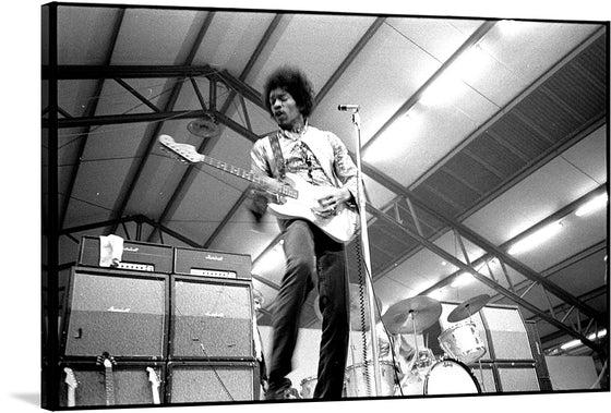 This black and white image captures a live performance scene of a musician playing an electric guitar. The musician’s face is obscured, but they wear a patterned shirt and dark pants and appear to be in motion. Several large amplifiers are visible behind the musician, and a drum set with cymbals is also seen in the background. 