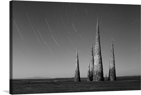“Subterrafuge Spires at AfrikaBurn in Monotone” is a mesmerizing artwork that captures the ethereal beauty of the AfrikaBurn event. The print features four towering spires standing tall against the backdrop of a star-streaked sky, captured in a stunning long exposure shot. The monotone palette enhances the haunting charm of the scene, creating a striking contrast between the textured spires and the smooth trails of stars. 